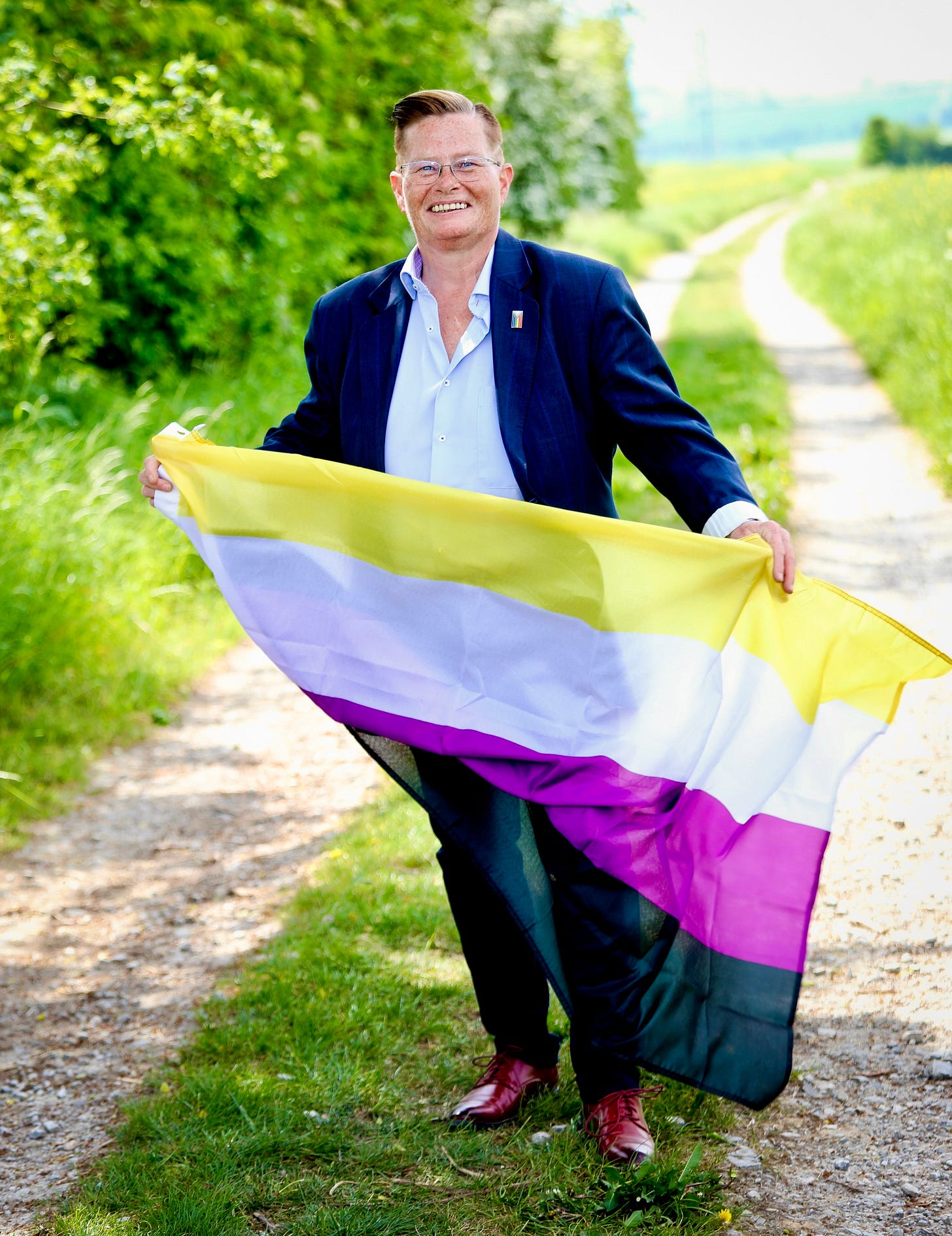 A person standing on a sunlit rural path surrounded by green fields, smiling and holding a flowing nonbinary pride flag with horizontal stripes of yellow, white, purple, and black. They wear a navy blazer, white shirt, dark pants, and red shoes.