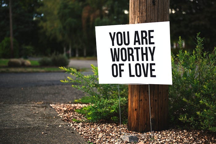 A white sign propped against a telephone pole. It reads: You are worthy of love. A white sign propped against a telephone pole. It reads: You are worthy of love.