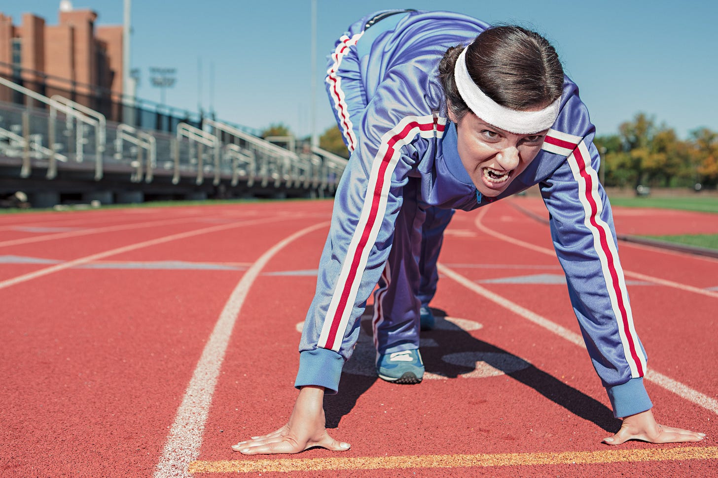 An athlete in a blue tracksuit and head band kneels at a starting line with a look of such fierce determination it approaches rage