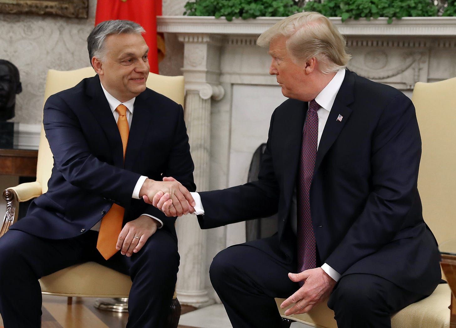 Two men shake hands in front of a fireplace. Man on left has salt-and-pepper hair and wears a yellow tie. Man on right has faded blonde hair and wears a purple tie. Two men shake hands in front of a fireplace. Man on left has salt-and-pepper hair and wears a yellow tie. Man on right has faded blonde hair and wears a purple tie.