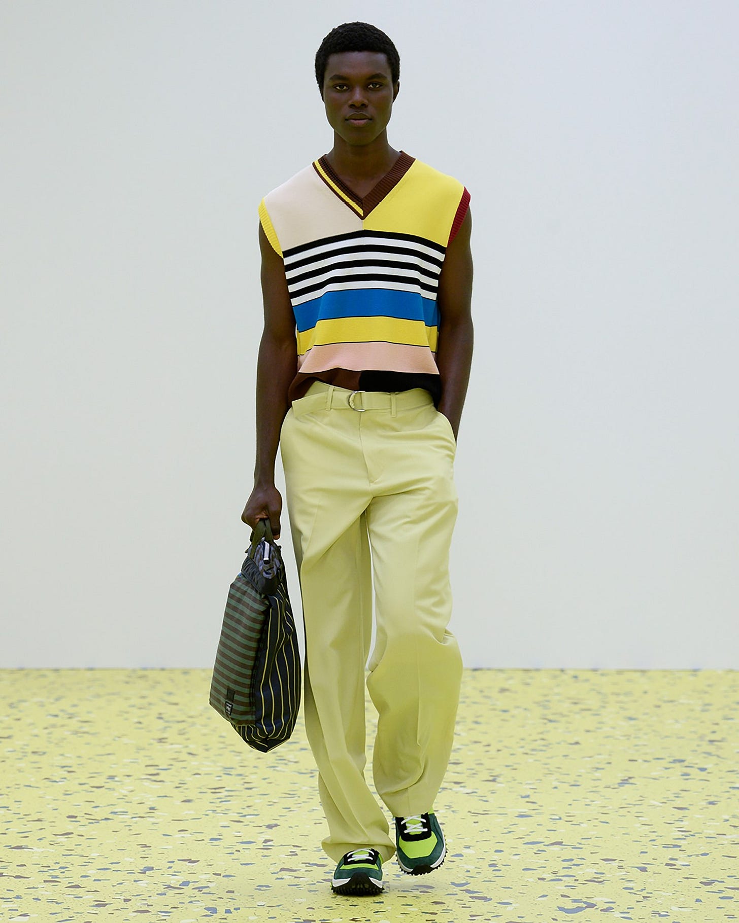 A young Black male model in a catwalk show who is wearing a sleeveless knitted vest with multicolour panels of yellow, brown and blue, complements the relaxed light yellow trousers, green and white sneakers and stripy tote he is carrying.