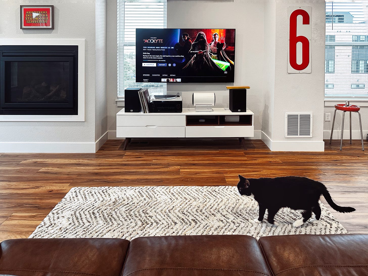 A photo of my living room with a couch, artwork on the walls, my cat walking across the carpet, and my media console with my TV hung on the wall, displaying the main screen of the show "The Acolyte" on Disney+.
