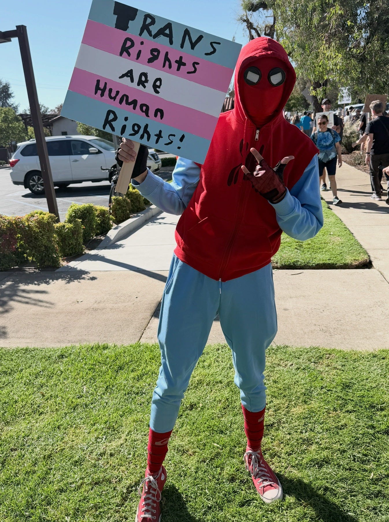 Someone in a spider-man costume holds up a protest sign that says "Trans Rights are Human Rights"