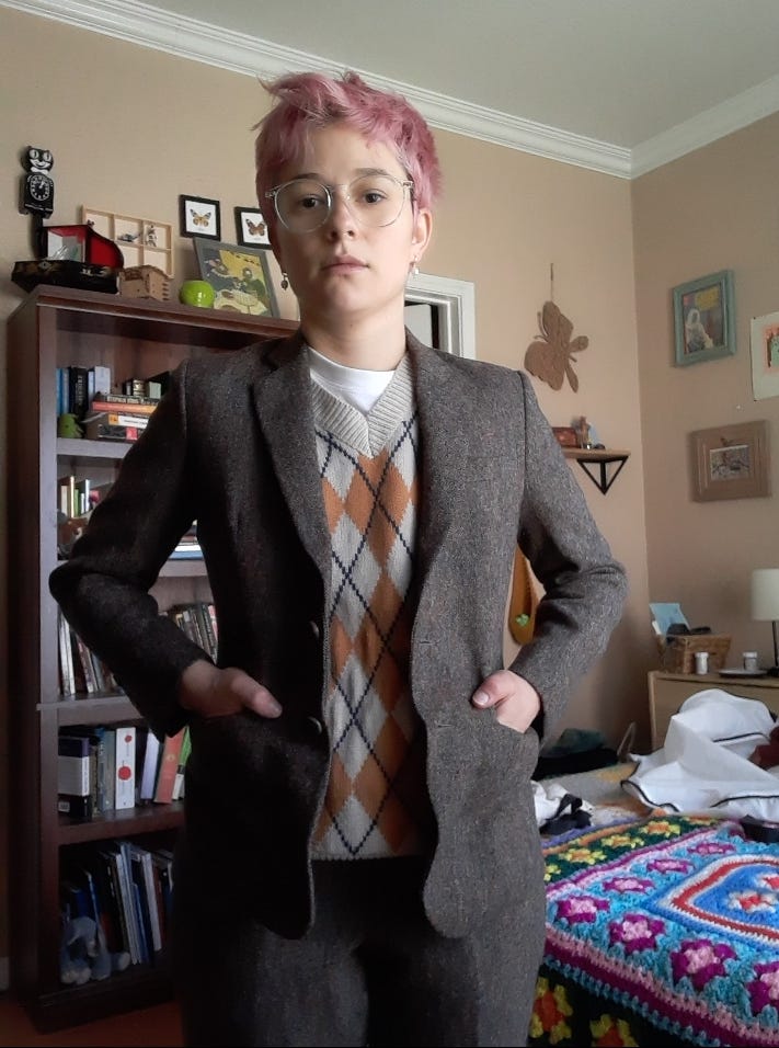 August stunting in a brown suit and tan-and-mustard sweater vest. They have pink hair and round glasses. Their hands are in their jacket pockets.