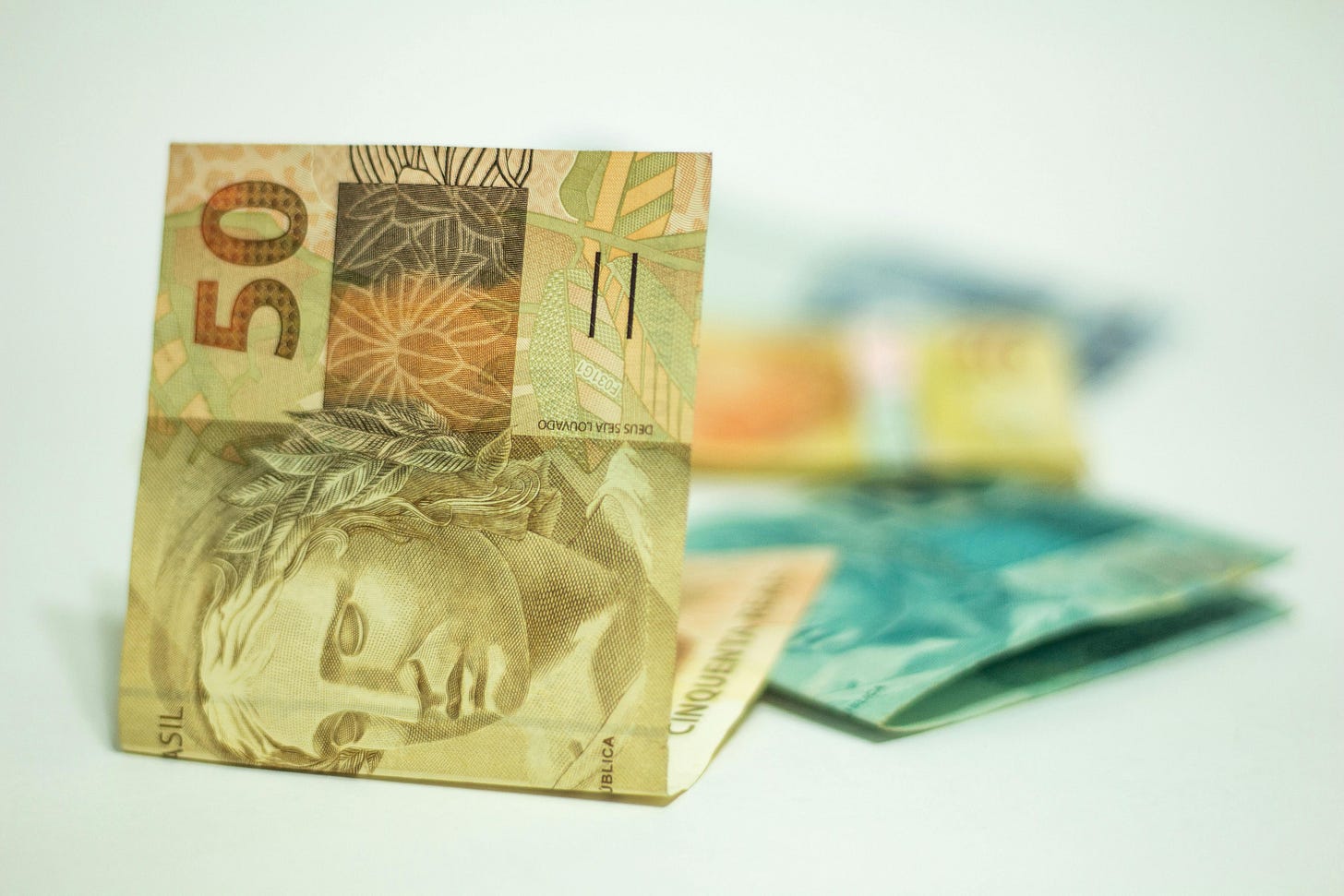 A imagem mostra várias cédulas de dinheiro brasileiro espalhadas sobre uma superfície clara. A nota mais próxima e em destaque é uma cédula de 50 reais, em tons de marrom e dourado. Nela aparece o rosto de uma figura feminina que representa a República, com detalhes finos e texturas típicas do real. Mais ao fundo, um pouco desfocadas, há outras notas de diferentes valores e cores — algumas azuladas, outras amareladas — sugerindo uma pequena pilha ou conjunto de cédulas sobre a mesa.
