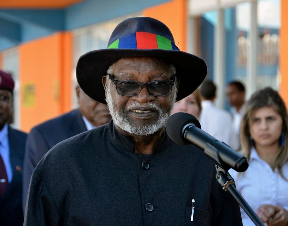 A man with a grey beard and a hat with a multicoloured band on it, in a black shirt and sunglasses, speaks into a microphone. A man with a grey beard and a hat with a multicoloured band on it, in a black shirt and sunglasses, speaks into a microphone.