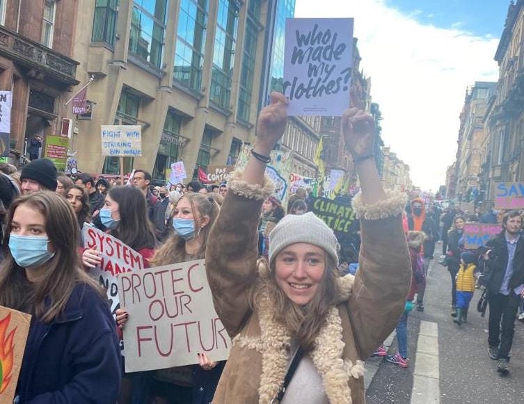 A picture of a young woman holding a sign that says 'Who made my clothes?' in a march in a street with tall old buildings. Crowds surround her.