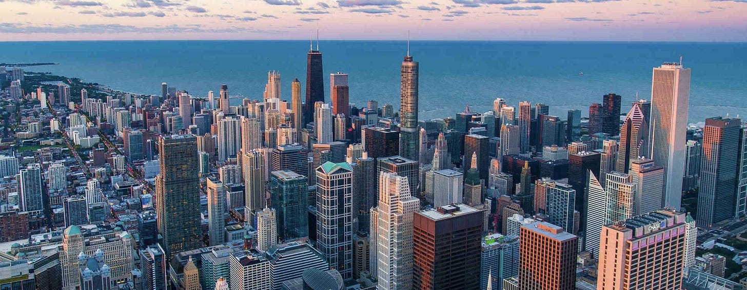 An aerial view of the Chicago skyline at dusk, featuring a mix of high-rise buildings and skyscrapers with Lake Michigan in the background. The sky is painted with soft hues of pink and blue, adding a serene backdrop to the bustling cityscape.