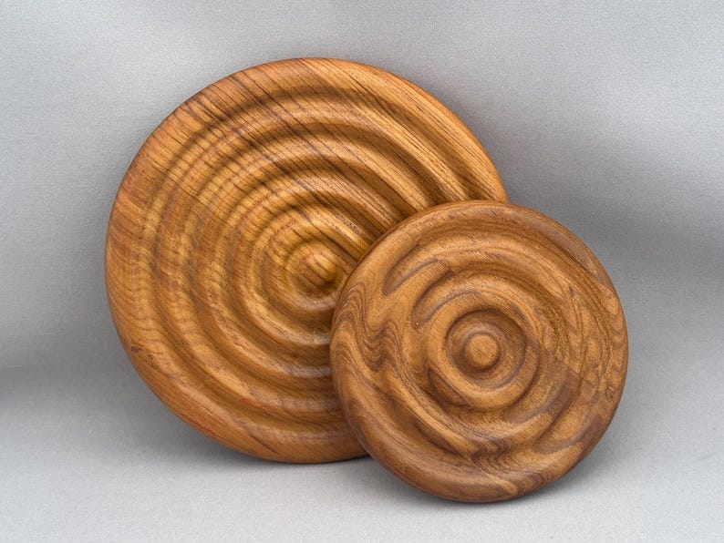 May include: Two round wooden coasters with concentric circles carved into the surface. The larger coaster is a light brown color and the smaller coaster is a darker brown color.