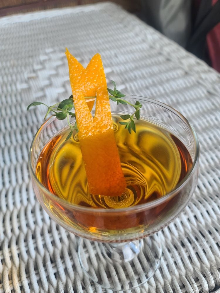 The Bijou cocktail in a coupe glass with orange lapel and thyme garnish sitting atop a wicker table.