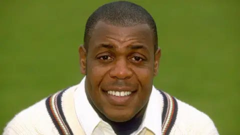 Getty Images David 'Syd' Lawrence. He is has brown eyes and short dark hair. He is pictured wearing a white cricket jumper with a stripe around the neck. He is looking at the camera and smiling. 