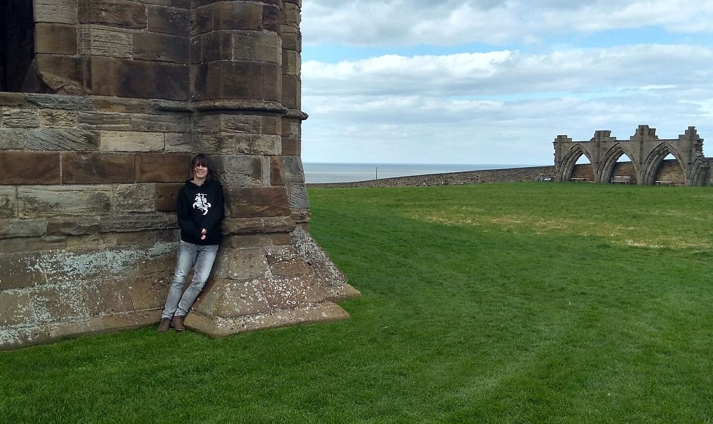 Picture of young woman in black hoodie and grey jeans leaning against old ruins on a cliff overlooking the sea
