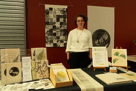 The table is covered in many different botanical and text based prints on different surfaces, including a fabric book. In the second photo I am standing at the stall, with two large fabric botanical print hangings on the wall behind me., In the first photo is a close up of the letterpress text test prints on calico. The text overlaps itself making a tall pillar of text overlapping and fading in and out of itself.