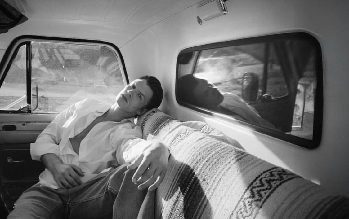 Black and white photograph of Connor Storrie lounging on the backseat of classic Porsche wearing jeans and a half-unbuttoned white button down and looking very cool.