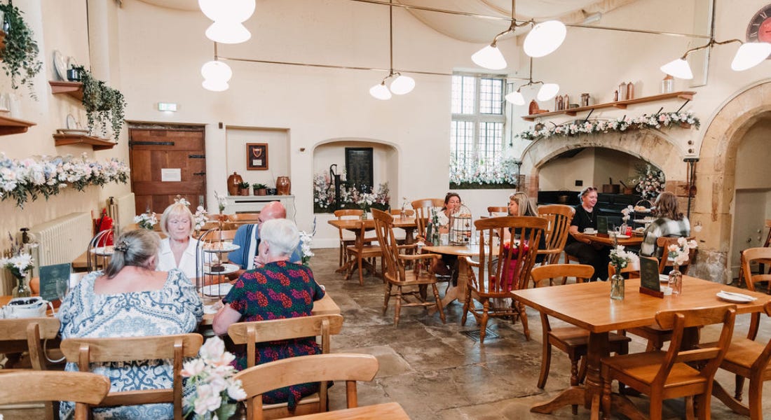 A tearoom with people sat at wooden tables, hanging pendant lights, and beige walls A tearoom with people sat at wooden tables, hanging pendant lights, and beige walls