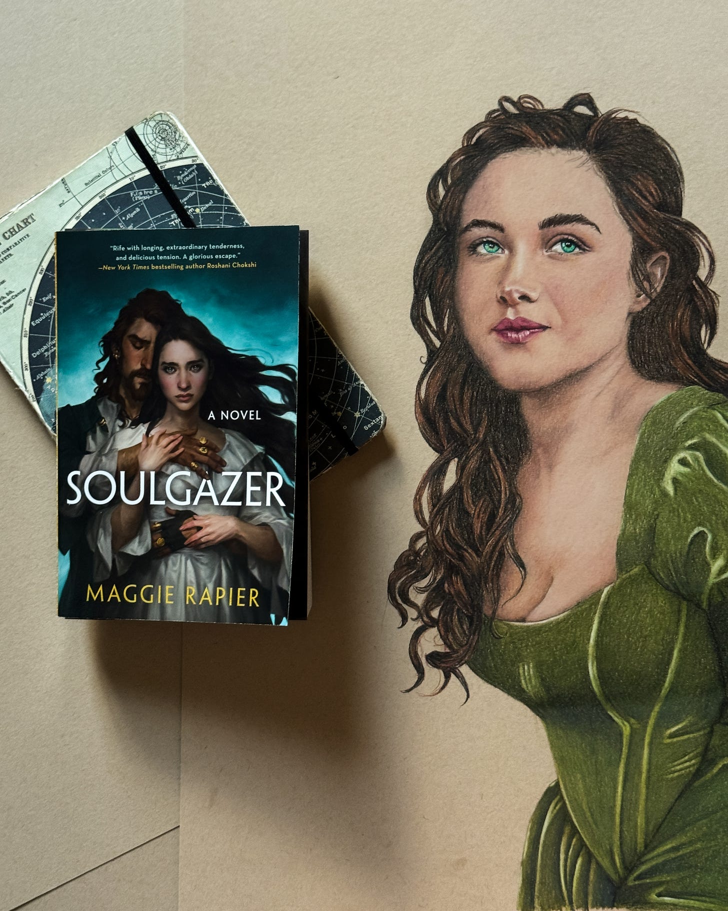 On a blank background, a portrait drawing of a curvy white woman with wavy, windswept brown hair and brilliant, ocean green-blue eyes rests next to a book titled SOULGAZER by Maggie Rapier, in which the same woman is painted on the cover standing in the arms of a white man with curly, reddish-brown hair wearing a green pirate coat, leather glove, several golden rings. The book is on top of a tattered journal with a navy blue chart of the starts. On a blank background, a portrait drawing of a curvy white woman with wavy, windswept brown hair and brilliant, ocean green-blue eyes rests next to a book titled SOULGAZER by Maggie Rapier, in which the same woman is painted on the cover standing in the arms of a white man with curly, reddish-brown hair wearing a green pirate coat, leather glove, several golden rings. The book is on top of a tattered journal with a navy blue chart of the starts.