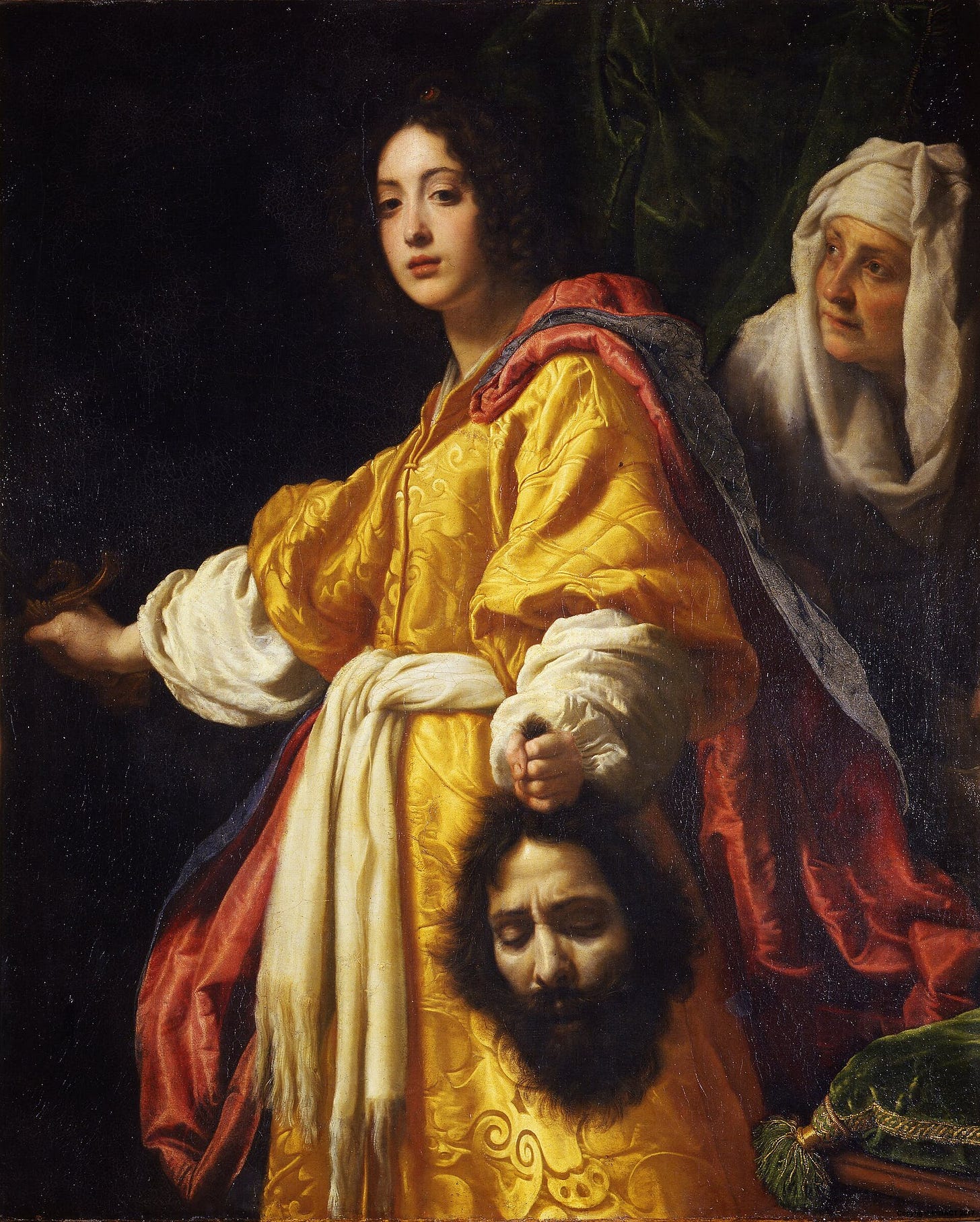 A young woman in a gold and blue brocade dress stands facing the viewer, holding a sword in one hand and a bearded man's severed head by the hair in the other, while an old woman in a white headscarf looks up at her from behind.