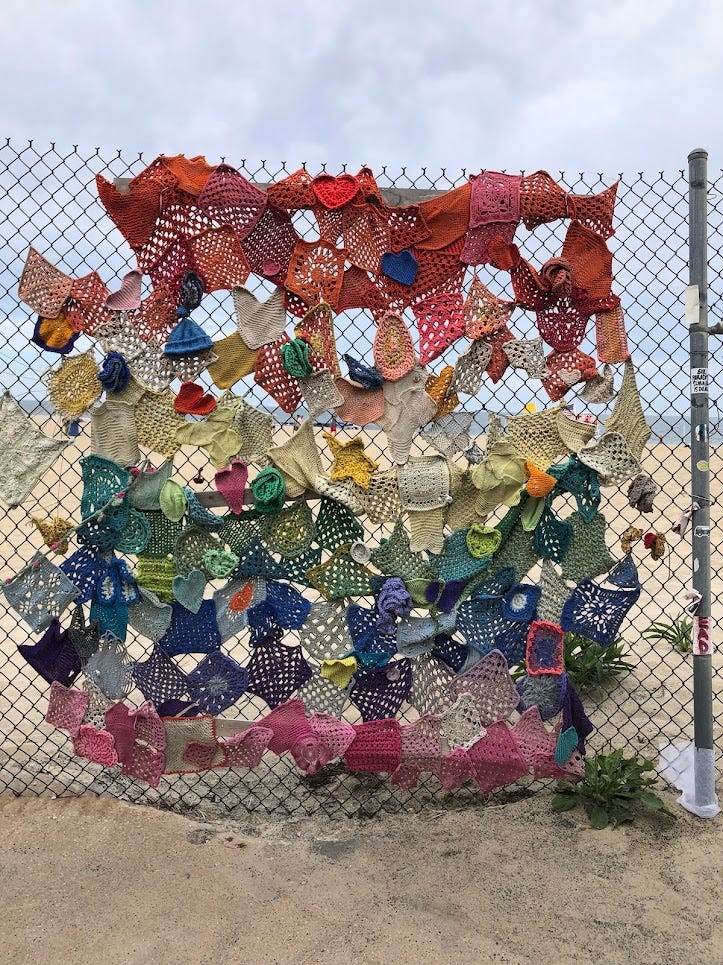 A piece of art built of many different crochet pieces, hanging on a chainlink fence in front of a beach. The pieces start red at the top, and slowly cycle through the colors of the rainbow, becoming purple at the bottom. A piece of art built of many different crochet pieces, hanging on a chainlink fence in front of a beach. The pieces start red at the top, and slowly cycle through the colors of the rainbow, becoming purple at the bottom.