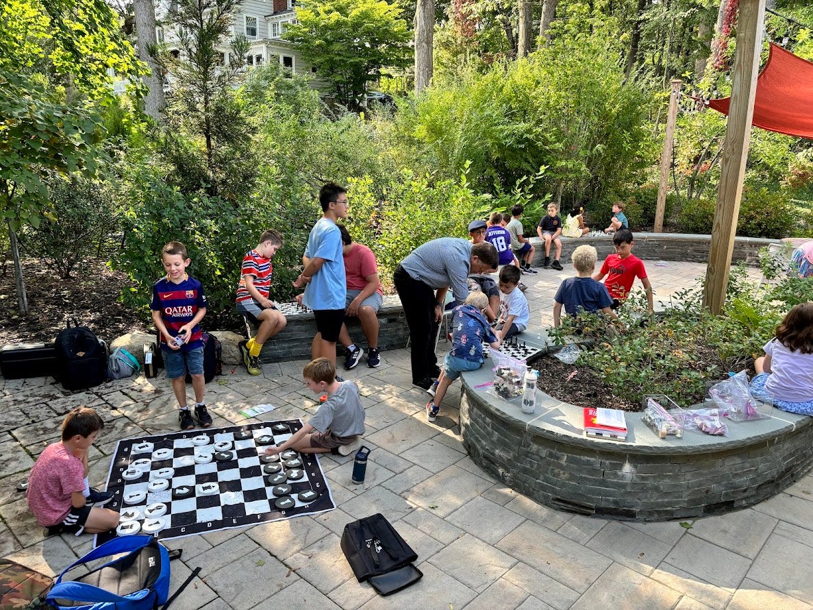 Chess around the ‘Bean’, the ML Library patio. Kids sitting on stone wall and the patio playing chess.