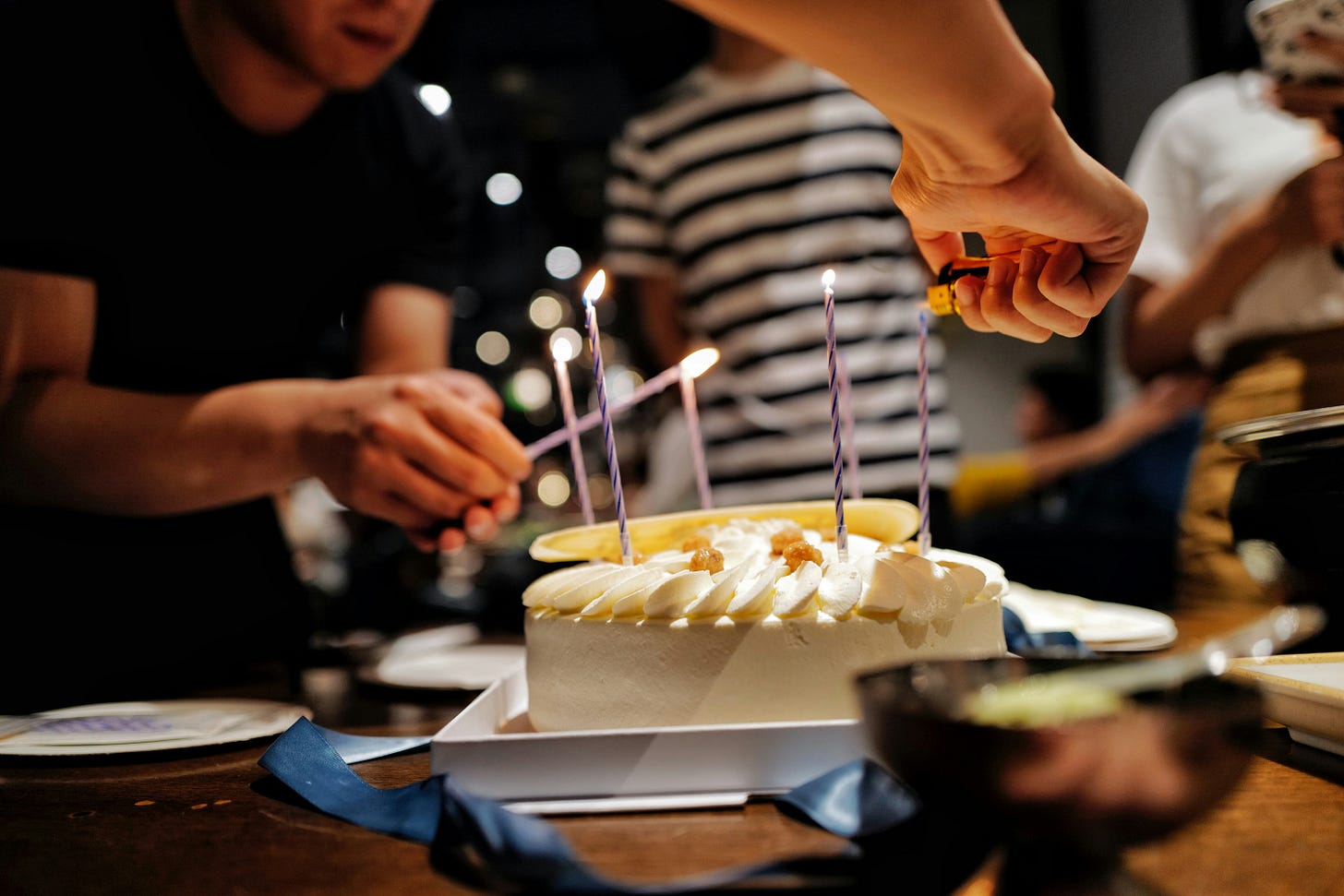 Pessoa acendendo velas de aniversário em um bolo branco com cobertura cremosa, enquanto outras observam ao fundo.