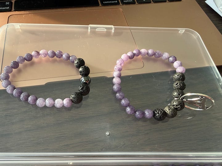Two bracelets with lavender colored stones and lava rock. One has a shiny bead and the other a yoga charm.