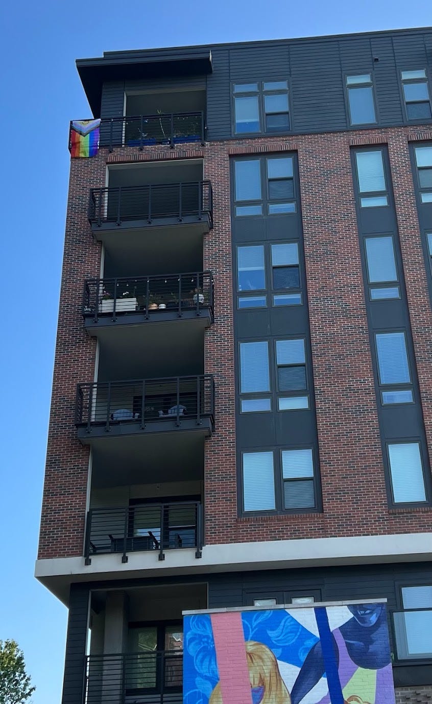 A brick apartment building against a blue sky. At the top is a balcony with a pride flag