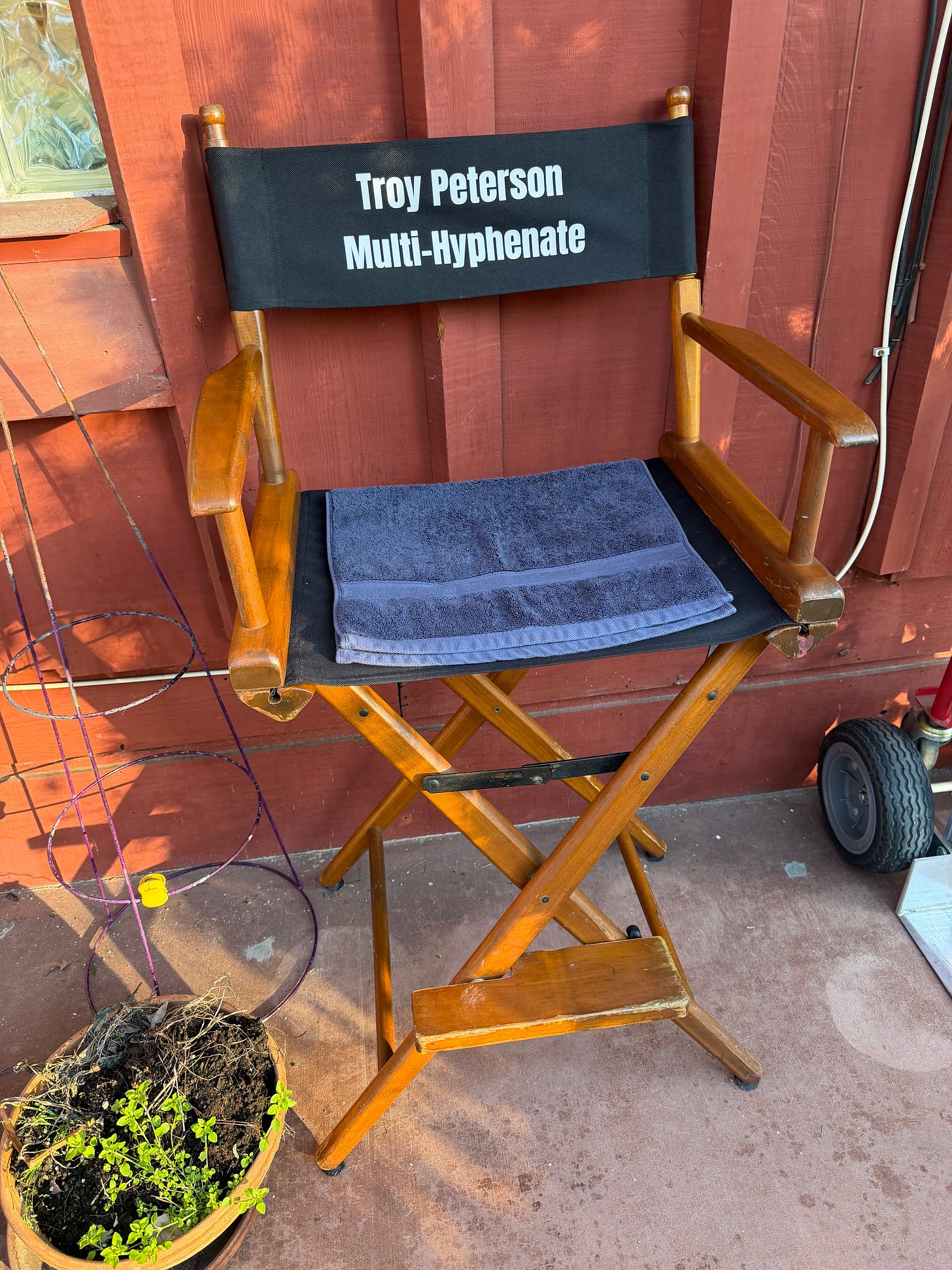 A wooden director’s chair sits against a red exterior wall, with black canvas reading “Troy Peterson Multi-Hyphenate.” A folded towel rests on the seat, and small set items, including a plant pot and equipment, are visible nearby.