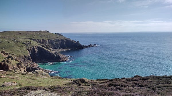 A photograph of a rugged coastal landscape of grass and rocks, looking down into a bay of very blue sea.