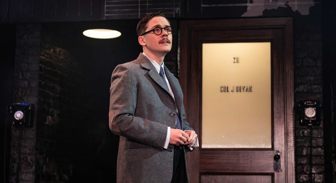 A man wearing a grey blazer and glasses, standing next to a wooden door on stage A man wearing a grey blazer and glasses, standing next to a wooden door on stage