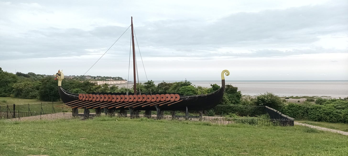 Replica of the Viking longship, The Hugin. A long thin boat wtih many oars and a tall mast. the sea and sky are visible behind