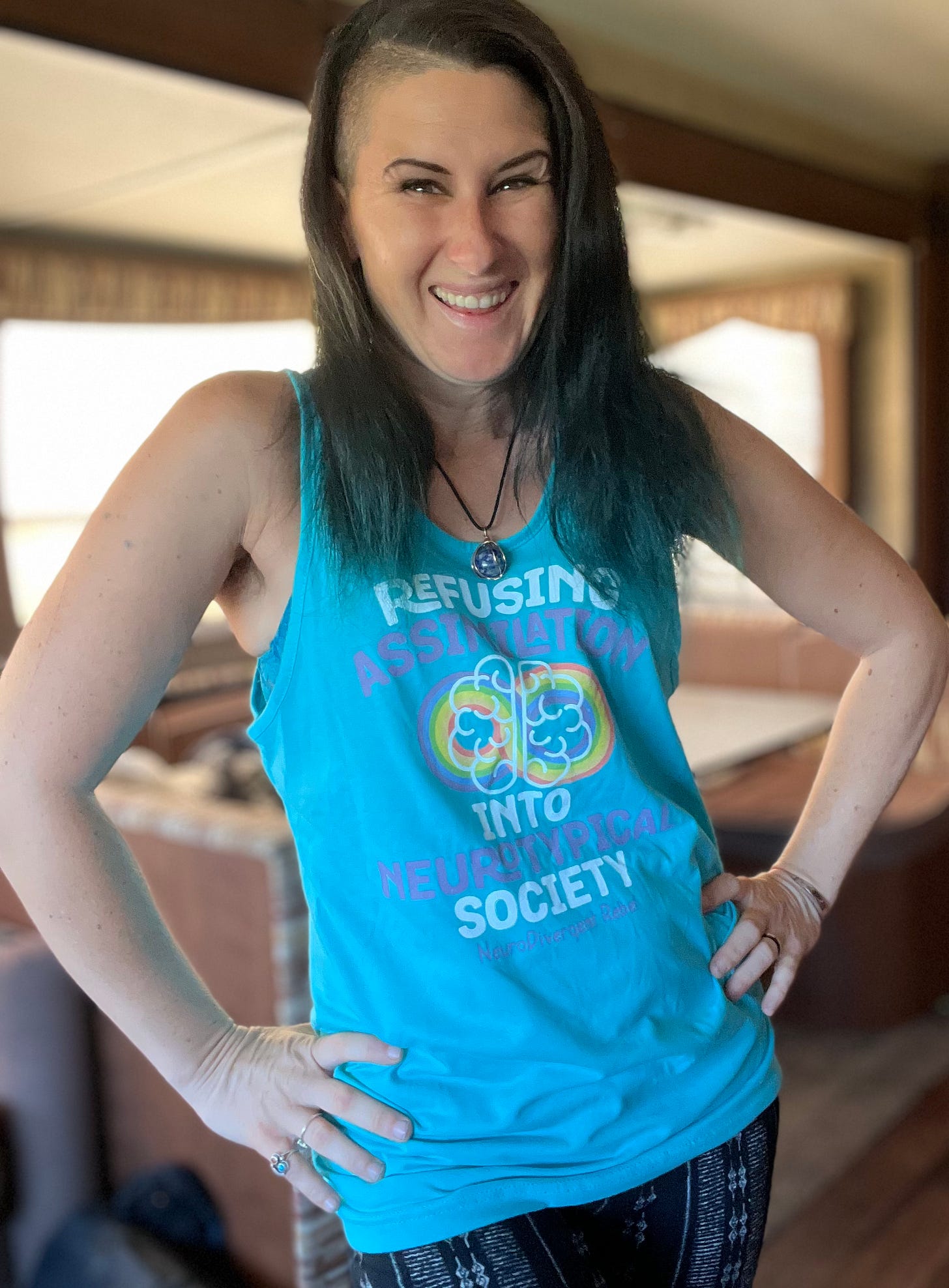 Lyric, poses happily in their RV wearing a teal tank top with a rainbow infinity on it that says “refusing assimilation into NeuroTypical society - NeuroDivergent Rebel” Lyric, poses happily in their RV wearing a teal tank top with a rainbow infinity on it that says “refusing assimilation into NeuroTypical society - NeuroDivergent Rebel”