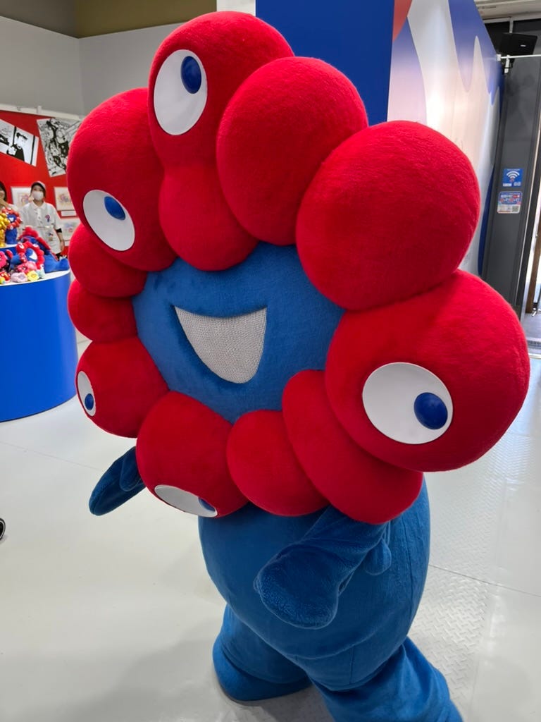 A person wearing a Myaku-Myaku costume. The blue face has a white smiling mouth and is surrounded by red blobs, 5 of which have eyes on them. The body is blue, and slightly rounded.