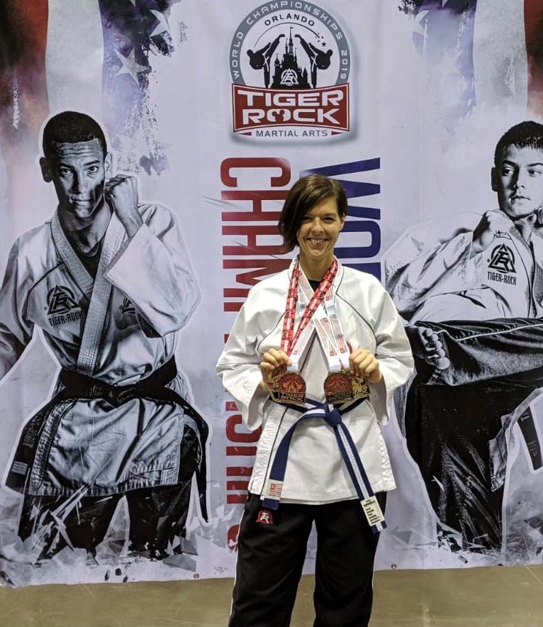 Smiling martial artist wearing a white uniform and blue belt stands holding two medals in front of a Tiger Rock Martial Arts championship backdrop featuring black-and-white images of competitors.