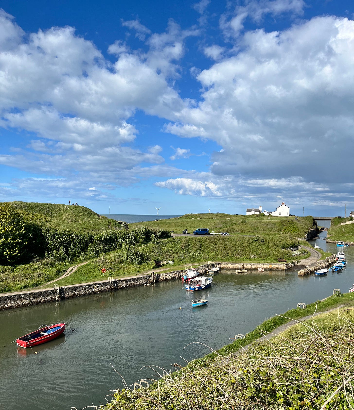 A view of Seaton Sluice harbour on a clear day, small boats on the water and a green coastal headland under a blue sky.