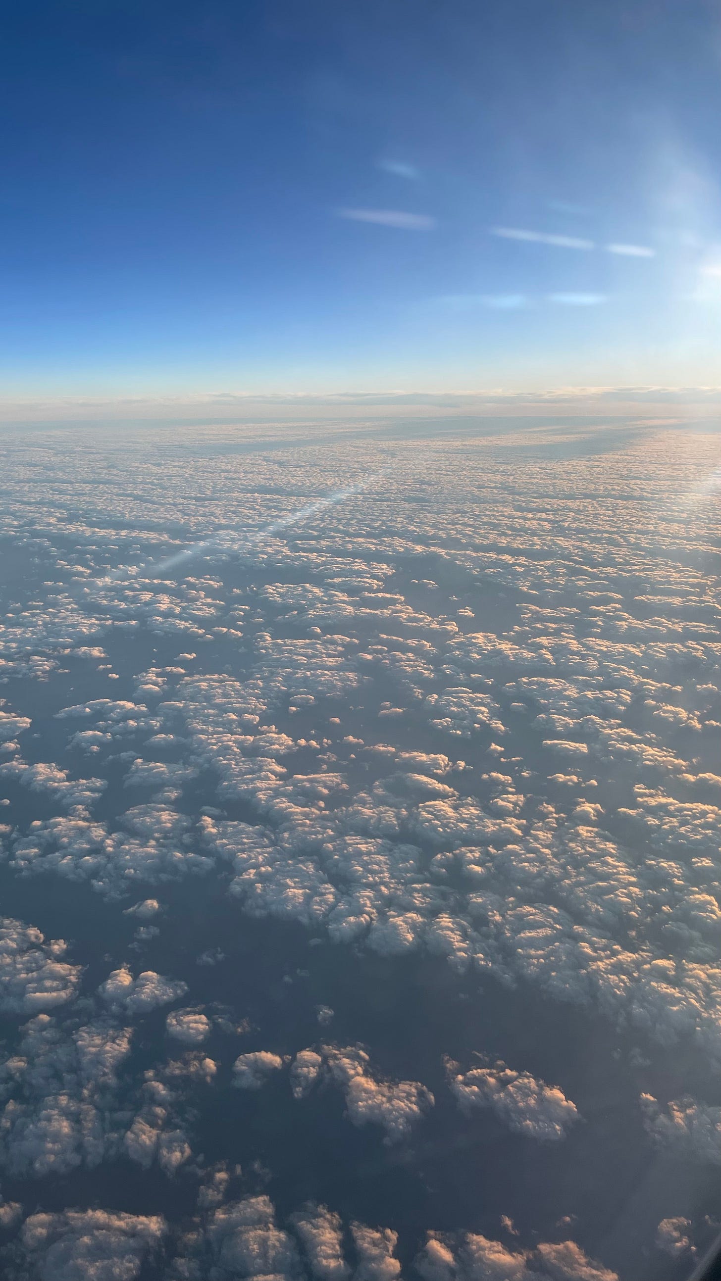 Rays of light over a field of puffy white and pink clouds as seen from above through the window of an airplane.