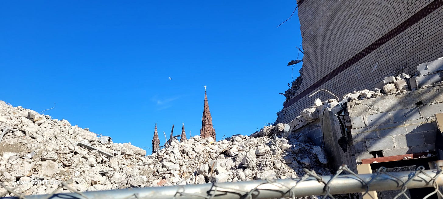 A large pile of gray refuse from a demolished building is in the foreground. Rising behind it is visible the top of a church, with a cross at the very top, a clear blue mid-day sky, and a faint moon.