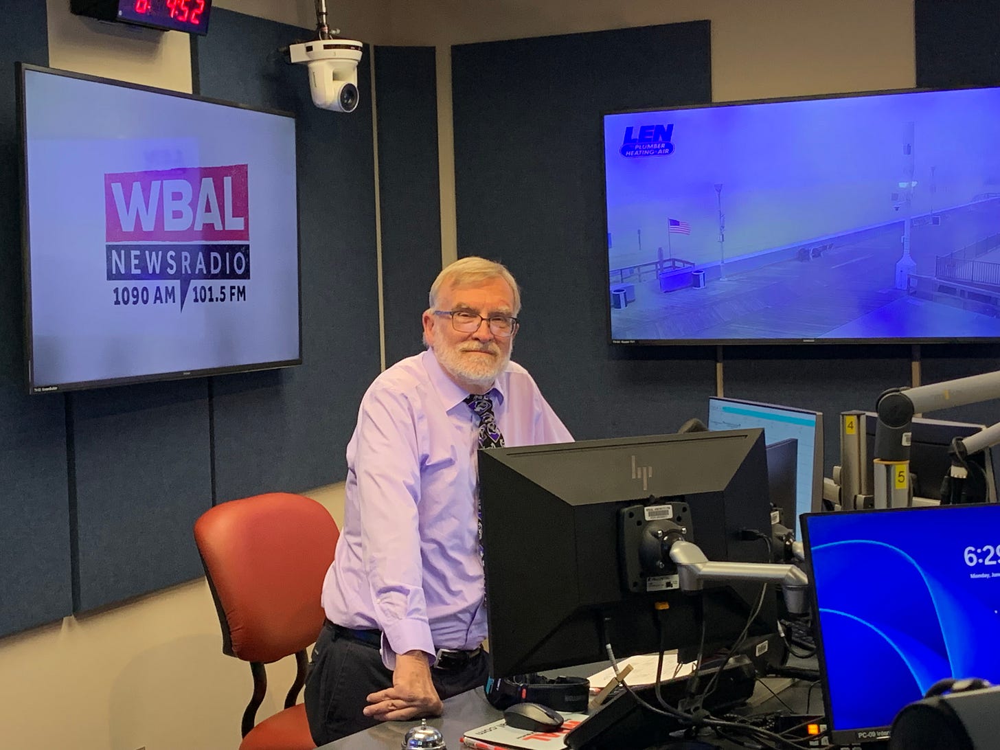 A man in a purple shirt and tie sits at a desk with a computer, identified as John Dedie at WBAL NEWSRADIO.