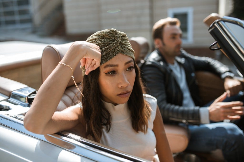 Woman facing outside in a convertible, looking sad. Man drives the convertible in foreground, looking like he’s talking, but she’s tuned-out. Woman facing outside in a convertible, looking sad. Man drives the convertible in foreground, looking like he’s talking, but she’s tuned-out.