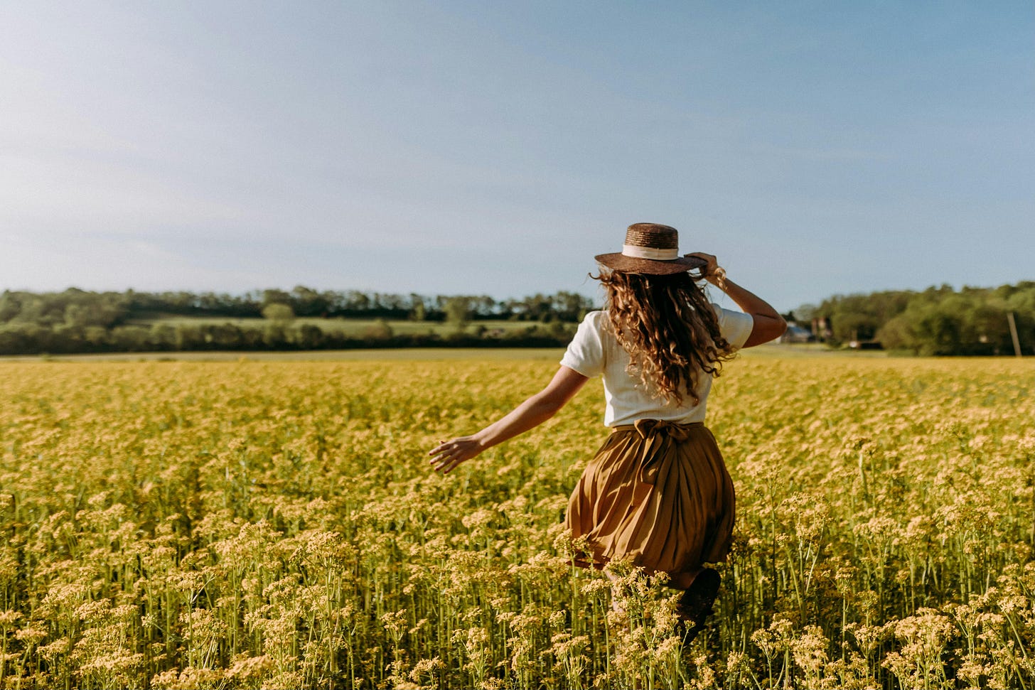 Back view of a woman with long hair wearing a brown skirt and hat running through a field of yellow flowers.