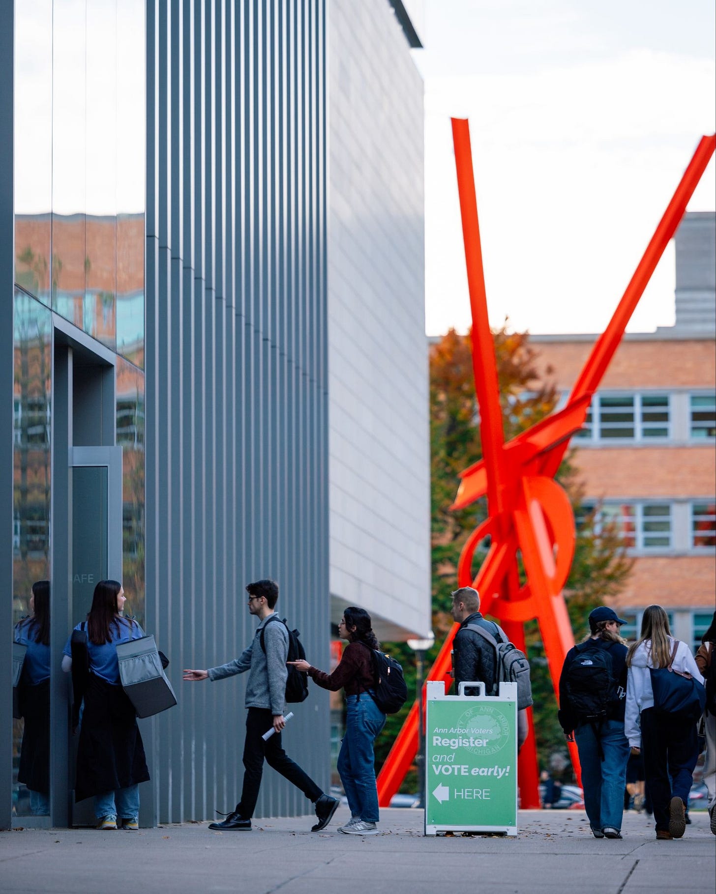A small line of students walk into the side entrance of UMMA near a green sign reading: “Ann Arbor Voters, Register and Vote Early Here”
