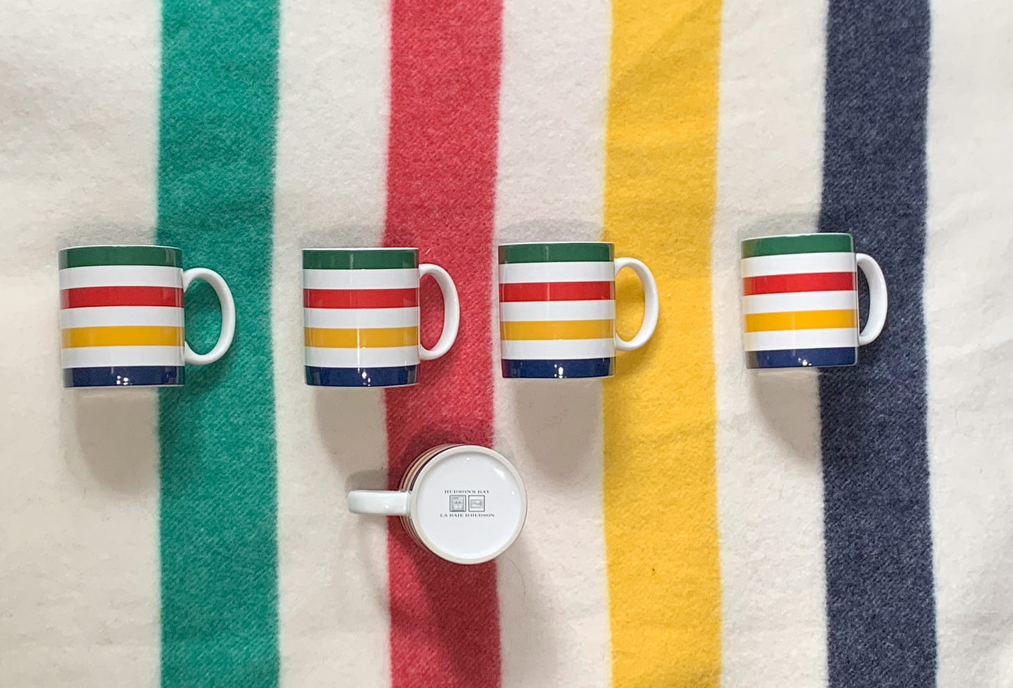 The classic Hudson’s Bay striped blanket. The stripes run: white, green, white, red, white, white, yellow, white, dark blue, white. On the blanket lie four similarly patterned mugs and below them, another upturned to display the bottom, with the Hudson's Bay logo and the name in both English and French (La Baie d'Hudson).