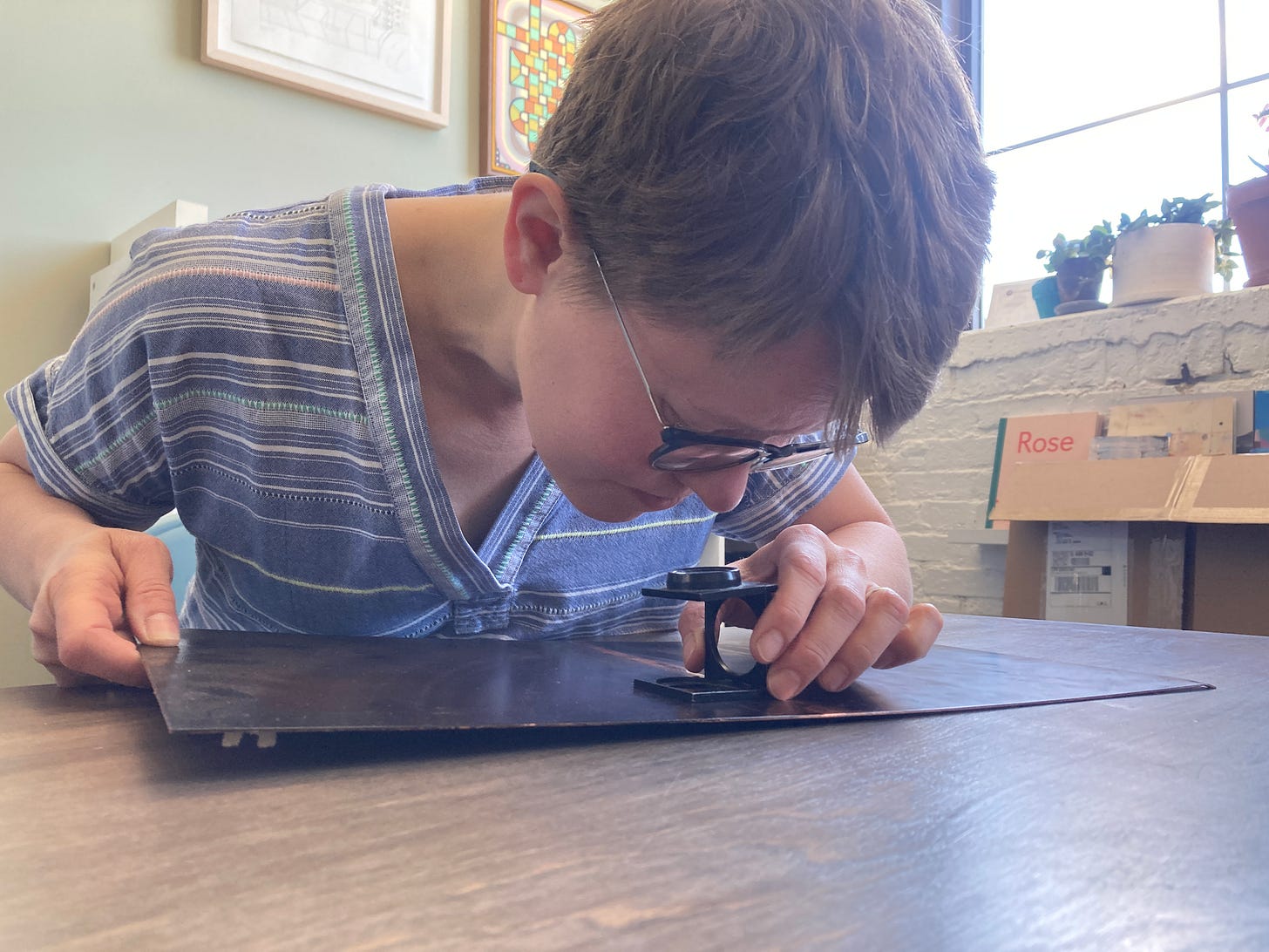 A white woman with short blonde hair and glasses leans over a loop resting on a metal plate. She is peering through the loop to closely examine the marks on the plate.