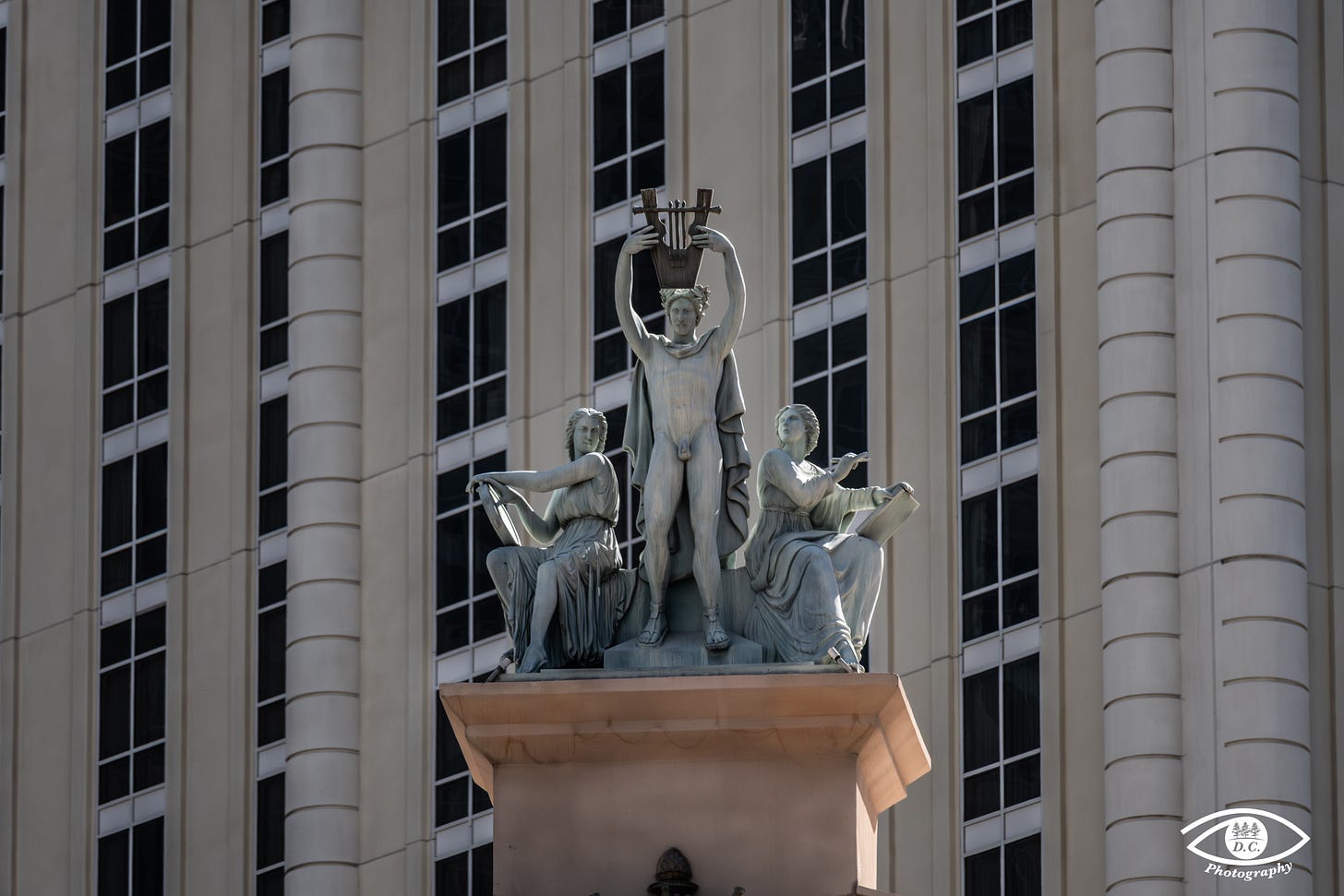A photograph of a monumental, classical-style statue group placed on a pedestal in front of a modern high-rise building. The central figure is a standing nude male holding a lyre above his head in both hands. He is flanked by two seated female figures in flowing robes. The statue is positioned against the background of a building facade with numerous vertical rows of identical, dark-paned windows alternating with light-colored, vertically-grooved columns or wall sections. The light is bright, casting some shadows.