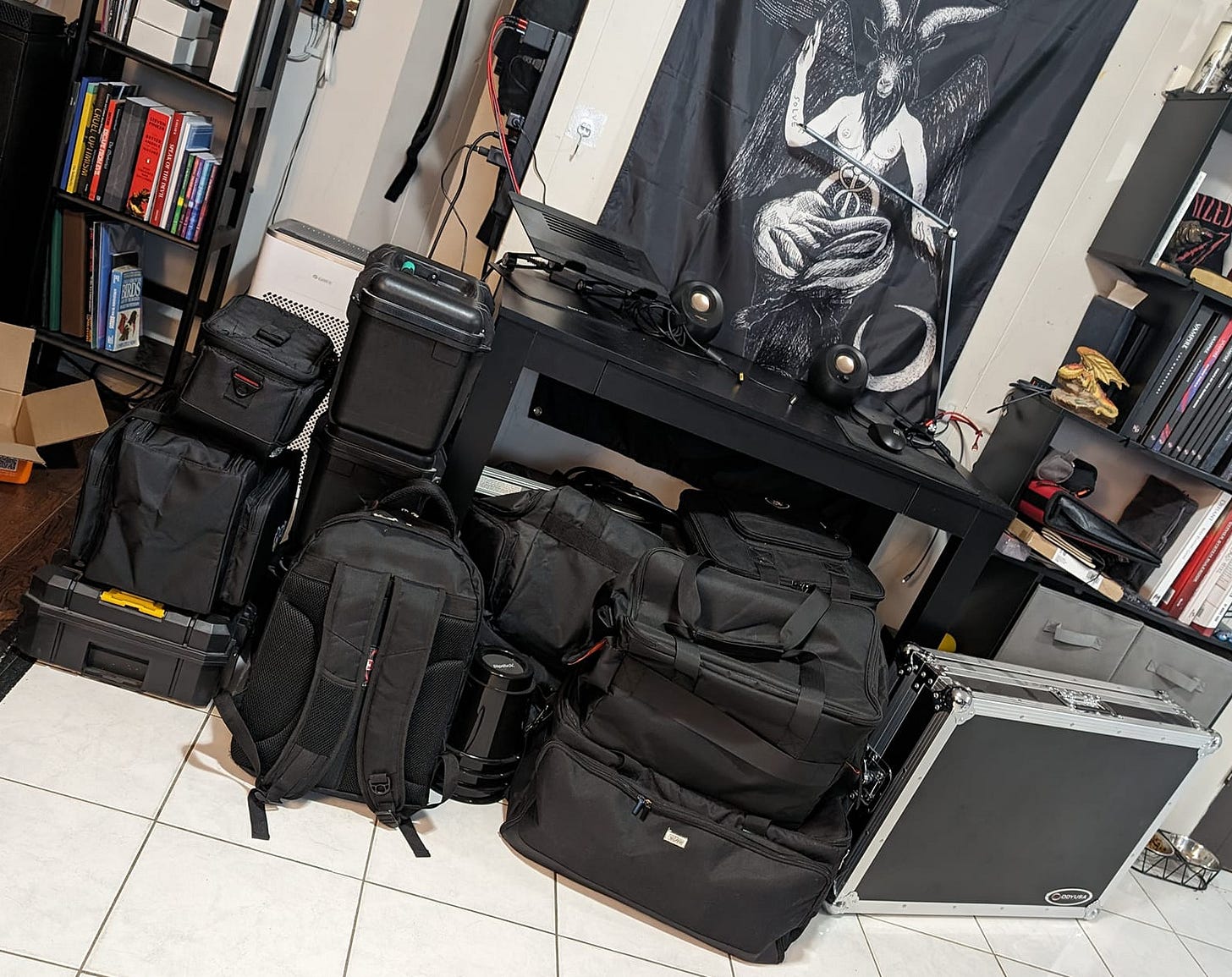 A large pile of gear bags sitting on a floor in front of two bookshelves, a desk, and a Baphomet flag