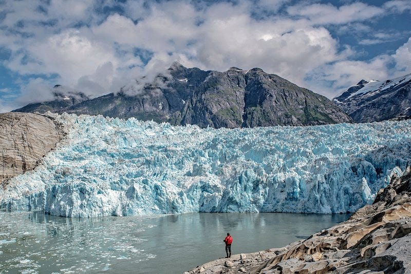 A photo of a person in a red jacket looking out at a melting glacier. There are mountains in the background.