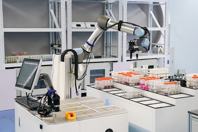 Robotic arm hovering over trays of specimen containers in a lab, a computer monitor behind it Robotic arm hovering over trays of specimen containers in a lab, a computer monitor behind it