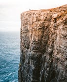 A tall, sheer cliff overlooking the sea with a tiny human figure on it.