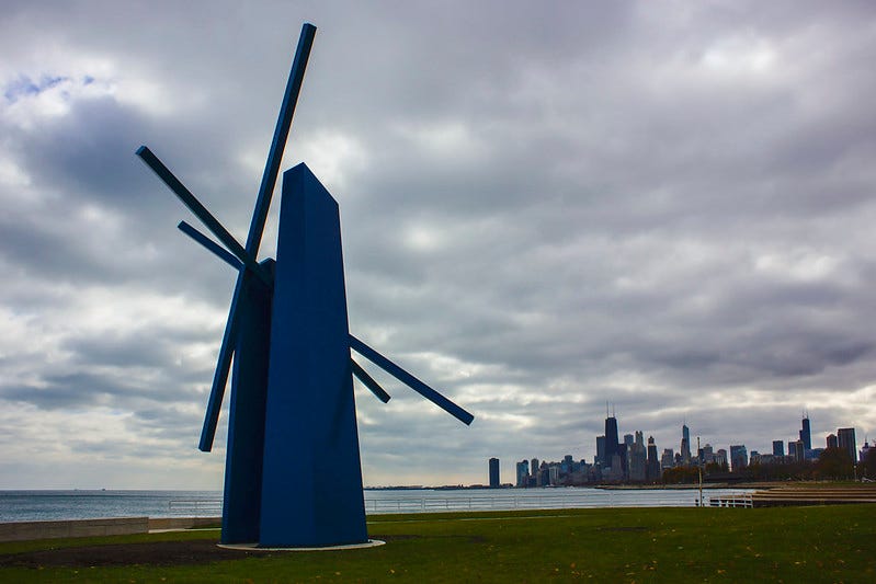 A large, abstract blue sculpture resembling a windmill stands in the foreground, with a cloudy sky and the Chicago skyline in the background. A large, abstract blue sculpture resembling a windmill stands in the foreground, with a cloudy sky and the Chicago skyline in the background.