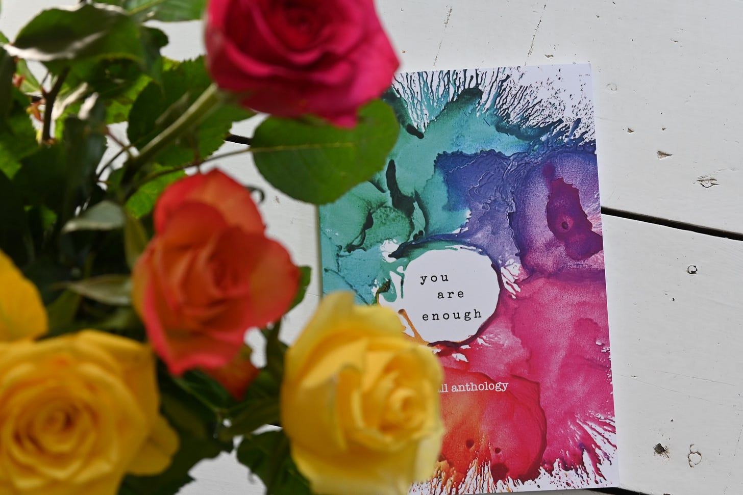 An image of anthology of essays called 'You are Enough' on a white wooden floor with a bouquet of roses in the foreground.