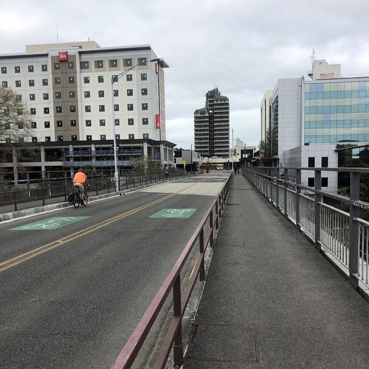 Left: A photograph from the footpath, looking west, showing footpath on both sides, with two lanes of traffic which doubles as sharrows. Right: The view from the Sonny Carpark footbridge, looking down onto the rail bridge, and then across and over onto Claudelands Bridge.